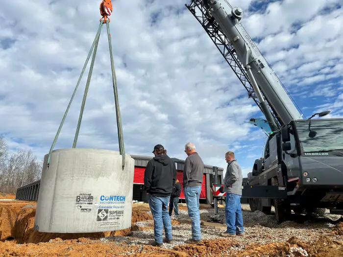 Construction crew guiding a crane lifting a large precast concrete stormwater system at an outdoor construction site.